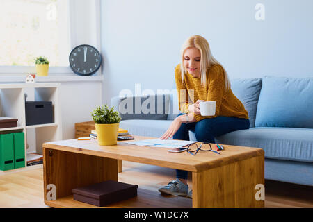 Schöne junge Frau sitzt auf einem Sofa zu Hause mit Kaffee und Dokumente Stockfoto