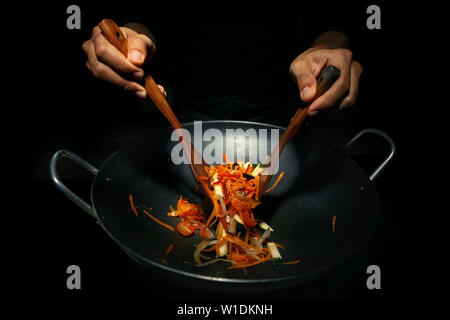 Hände des arbeitenden Küchenchefs, der eine Nudel mit Tomatenzucchini und Karotte im Wok in der Dunkelheit vorbereitet. Traditionelle Art der Kochplatte thai auf Wok Bratpfanne. Stockfoto