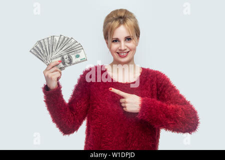 Gerne fröhliche Schöne junge geschäftsfrau in rot Bluse, die hielten viele Dollar, Zeigefinger und Kamera mit toothy Lächeln. In Stockfoto