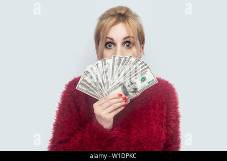 Portrait von Reichen attraktiven jungen Erwachsenen Geschäftsfrau in rot Bluse stehen und zeigen Fan von Bargeld Dollar mit großen verrückt Augen. Von der Kamera suchen, ICH Stockfoto