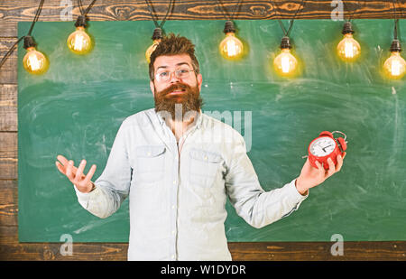 Zeitplan und Regime Konzept. Mann mit Bart und Schnurrbart auf verwirrten Gesichtsausdruck im Klassenzimmer. Bärtige hipster hält, schwarzes Brett für den Hintergrund. Lehrer in Brillen hält Wecker. Stockfoto