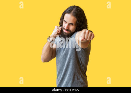 Portrait von Angry starke bärtiger junger Mann mit langen, lockigen Haar grau tshirt stehend mit Boxing Fäuste, suchen und Stanzen an Kamera. indoor Stud Stockfoto