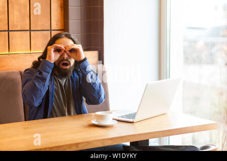 Portrait von Spionage hübscher junger erwachsener Mann Freiberufler in lässigen Stil im Cafe sitzen mit Laptop, die Hand in der Nähe von Augen, Fernglas mit Hand Stockfoto