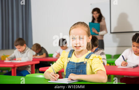 Niedliche intelligente jugendliche Mädchen studieren im Klassenzimmer auf Hintergrund mit Klassenkameraden und Lehrer Stockfoto