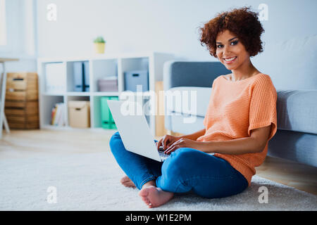 Schöne afrikanische Frau sitzt auf dem Boden mit dem Laptop zu Hause Stockfoto