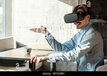 Portrait von Happy positive bärtiger junger erwachsener Mann in Blue Denim Shirt sitzen, vr und Video am Simulator mit erhobenen Hand und holdin Stockfoto