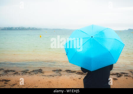 Zurück oder hinten Blick auf eine Frau, die durch das Meer in Regenjacke und mit einem blauen Schirm an einem regnerischen und Moody Tag. Wetter Konzept mit kopieren. Oran Stockfoto