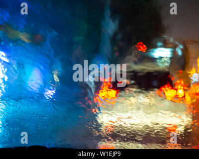 Verschwommen Regentropfen auf dem Auto Fenster mit Straßenbeleuchtung bokeh an regnerischen Jahreszeit Zusammenfassung Hintergrund. Wassertropfen Textur auf das Glas von dem Regen im Auto drivin Stockfoto