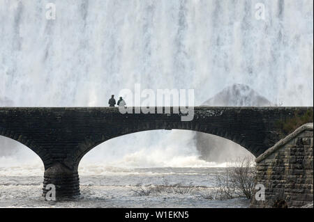 Die Besucher gehen über die Brücke wie Wasserkaskaden über Caban-coch Damm, bei Elan Valley Village in der Nähe von Rhayader in Powys, Wales, Vereinigtes Königreich, Europa Stockfoto