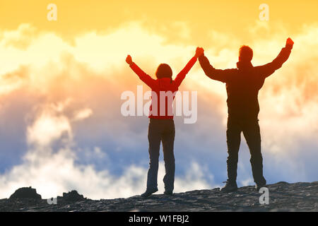 Glückliche Gewinner erreichen Leben Ziel - Erfolg Menschen am Gipfel. Business Achievement Konzept. Zwei Paar zusammen die Arme in die Luft des Glücks mit Vollendung in den Wolken bei Sonnenuntergang. Stockfoto