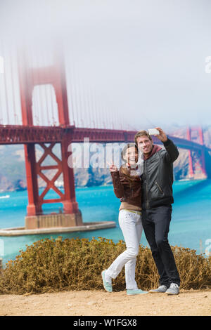 Touristen Paar unter selfie Foto in San Francisco die Golden Gate Bridge. Interracial junge moderne Paar mittels Smart Phone von berühmten amerikanischen Wahrzeichen. Asiatische Frau, kaukasische Mann. Stockfoto