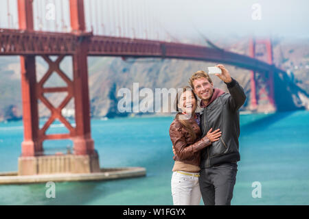 Glückliches junges paar Touristen unter selfie in San Francisco die Golden Gate Bridge. Interracial junge moderne Paar mittels Smart Phone. Asiatische Frau, kaukasische Mann. Stockfoto