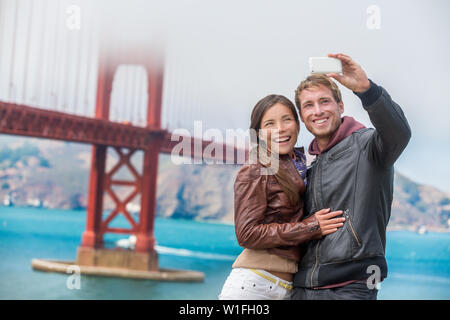 Paar Touristen unter selfie Foto in San Francisco die Golden Gate Bridge. Interracial junge moderne Paar mittels Smart Phone von berühmten amerikanischen Wahrzeichen. Asiatische Frau, kaukasische Mann. Stockfoto