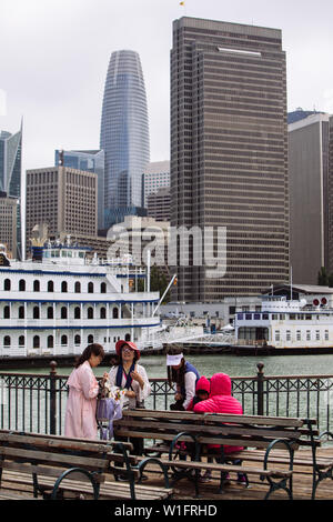 Asiatische Touristen ruhen sich auf dem hölzernen Pier 41 mit der San Francisco Belle Wheeler Charter Yacht und dem San Francisco Financial District auf der Rückseite aus Stockfoto