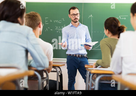 Junger Mann Lehrer mit Buch gibt interessante Vortrag für Schüler während der Lektion im Klassenzimmer Stockfoto