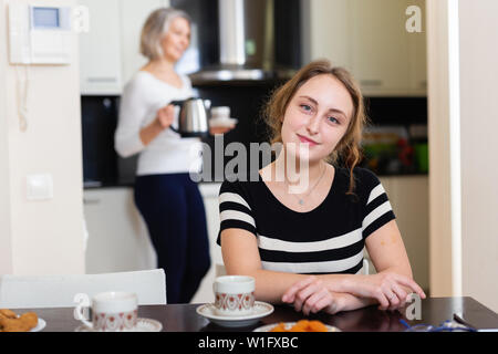 Lächelnden jungen Frau genießen Kaffee am Küchentisch, ältere Mutter im Hintergrund Stockfoto