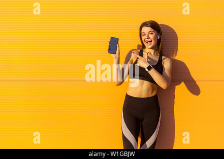 Junge zufrieden schöne sportliche Frau in Schwarz Sportbekleidung steht in der Nähe von Orange Wand Hintergrund und halten Telefon, Zeigefinger mit geöffnetem Mund, l Stockfoto