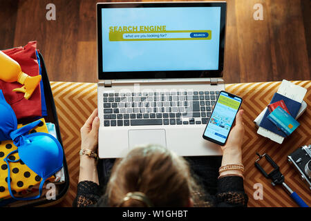 Junge Frau in Schwarz Bluse sitzen auf der Couch in der modernen Wohnzimmer mit Online Travel Hotel Reservierung site auf einem Smartphone und mit Search Engine Stockfoto