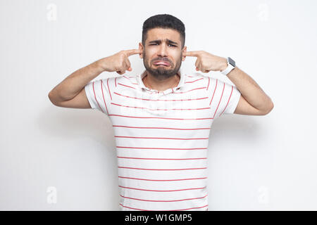 Ich will das nicht hören. Portrait von traurig verwirrt bärtiger junger Mann in gestreiften T-Shirt stehend, auf Kamera und legte seinen Finger auf die Pflege von Ohren. Stockfoto