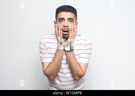 Portrait von schockiert gut aussehender bärtiger junger Mann in gestreiften T-Shirt auf Gesicht und Kamera mit unglaublichen Gesichtsausdruck. Stockfoto