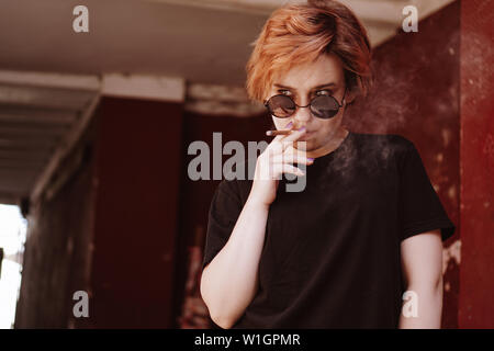 Tausendjährige cool hübsches Mädchen mit kurzen roten Haaren und Spiegel Sonnenbrille rauchen Zigarette in die alte Stadt mit roten Wänden Stockfoto