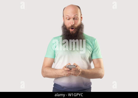 Portrait von schockiert im mittleren Alter bärtiger Mann mit Glatze bei smart phone Display mit überraschten Gesicht. Lesen gute Nachrichten oder etwas Erstaunliches. Stockfoto