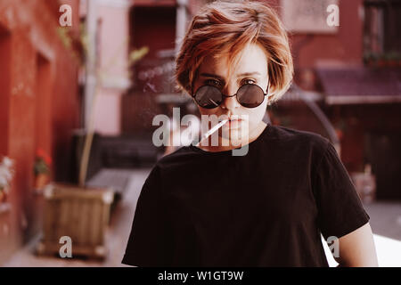 Tausendjährige cool hübsches Mädchen mit kurzen roten Haaren und Spiegel Sonnenbrille rauchen Zigarette in die alte Stadt mit roten Wänden Stockfoto