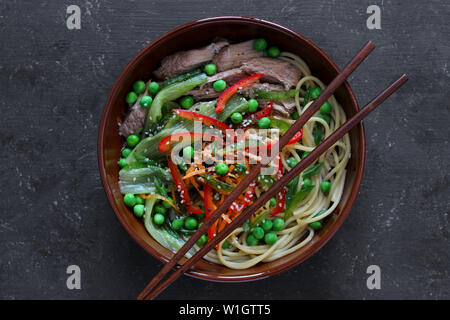 Nudeln mit Rindfleisch und Gemüse in Schüssel auf dunklem Hintergrund. Ansicht von oben mit der Kopie. Asiatische Küche. Stockfoto