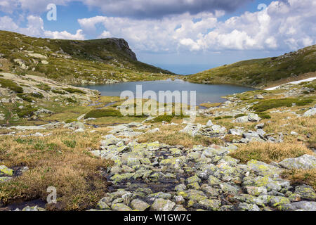 Vordergrund Felsen bedeckt durch gelbe Moos, blauer See, felsigen Gipfel und erstaunliche sonnenbeschienenen Landschaft von Rila Gebirge in Bulgarien Stockfoto