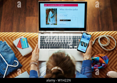 Obere Ansicht der elegante Frau in Blau Bluse sitzen auf der Couch in der modernen Wohnzimmer shopping Kleidung im Online Store auf einen Laptop und Smartphone. Stockfoto