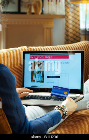 Junge Frau mit Kreditkarte surfen Online Fashion Retail ecommerce Aufstellungsort während auf Diwan im modernen Haus im sonnigen heißen Sommertagen sitzen. Stockfoto