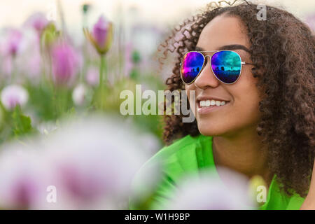 Gemischte Rasse schöne Afrikanische Amerikanische biracial Mädchen Teenager weibliche junge Frau trägt blaue aviator Sonnenbrille im Bereich der rosa Blumen Mohnblumen bei Su Stockfoto