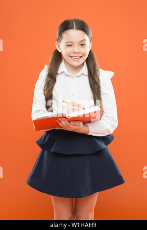 Schulmädchen Notizen auf orange Hintergrund. kleine Mädchen in Schuluniform. Schulmädchen holding Lektion buchen. Angaben form Buch. zurück zu Schule. nettes Mädchen halten Sie Notepad oder Tagebuch. Tagebuch für Mädchen. Stockfoto