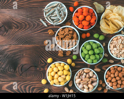 Sortiment von verschiedenen Snack für Bier, Wein, Party. Erdnüsse in Coconut Glasur, grüne vasabi, rote scharfe Chili, gelben Käse Glasur, Chips, Pistazie, Cracker, Fisch auf braune Holztisch. Platz kopieren Stockfoto