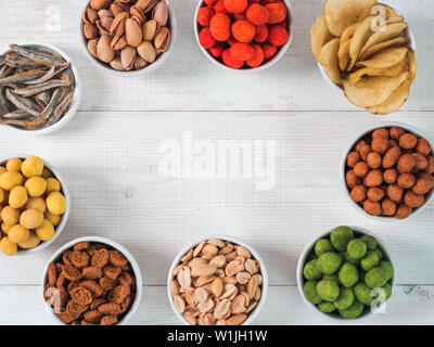 Sortiment von verschiedenen Snack für Bier, Wein, Party. Erdnüsse in Coconut Glasur, grüne vasabi, rote scharfe Chili, gelben Käse Glasur, Chips, Pistazie, Cracker, Fisch auf weiße Holztisch. Platz kopieren Stockfoto