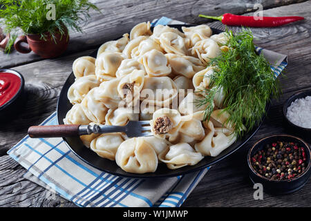 Nahaufnahme der hausgemachte heiße Teigtaschen mit Hackfleisch Füllung auf eine schwarze Platte mit frischem Dill und Tomatensauce in eine Schüssel auf einen hölzernen Tisch serviert, Hori Stockfoto