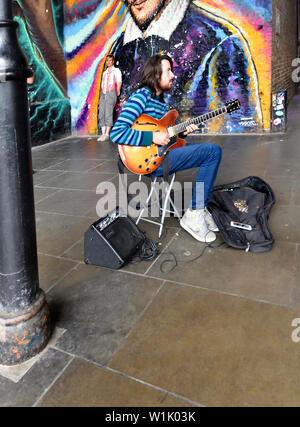 London, England, UK. Gaukler spielt die Gitarre vor ein Wandbild in Southwark - Menschen ein Foto im Hintergrund Stockfoto
