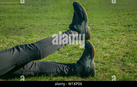 Die Beine des Athleten in Leggings und Sneakers, auf dem Boden liegend auf der Wiese in der Nähe. gekreuzten Beinen. Ausbildung auf dem freien. Warm-up, Stretching. Stockfoto