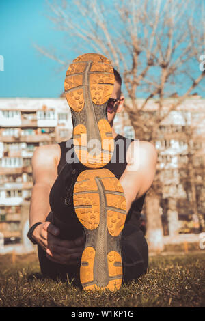 Die Beine des Athleten in Leggings und Sneakers, auf dem Boden liegend auf der Wiese in der Nähe. gekreuzten Beinen. Ausbildung auf dem freien. Warm-up, Stretching. Stockfoto