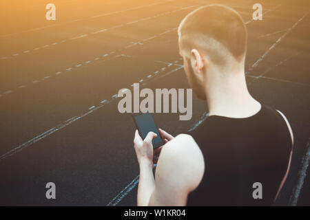 Ein junger Mann in schwarzer Kleidung am Telefon sucht. fitness Athleten auf dem Sportplatz. Training mit Gadgets. Warm up Körper Vorbereitung für die Summe Stockfoto