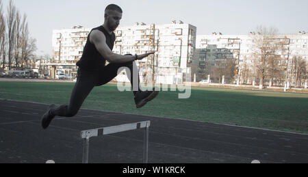 Ein Kaukasier Männlich in einem Sprung über ein Hindernis. läuft auf das Stadion. Leichtathletik Läufer in Sport einheitliche im Flug. energetischen körperlichen Aktivitäten. Stockfoto