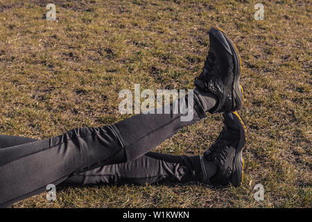 Die Beine des Athleten in Leggings und Sneakers, auf dem Boden liegend auf der Wiese in der Nähe. gekreuzten Beinen. Ausbildung auf dem freien. Warm-up, Stretching. Stockfoto