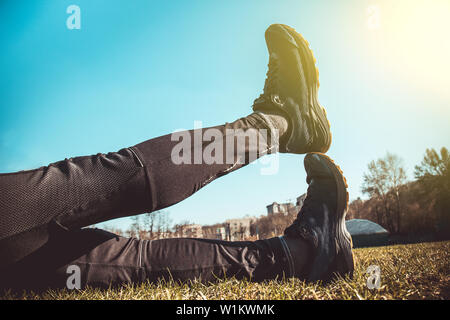 Die Beine des Athleten in Leggings und Sneakers, auf dem Boden liegend auf der Wiese in der Nähe. gekreuzten Beinen. Ausbildung auf dem freien. Warm-up, Stretching. Stockfoto