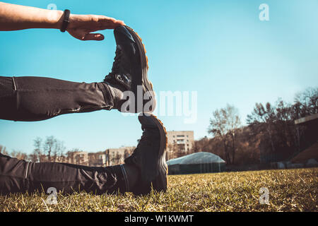 Die Beine des Athleten in Leggings und Sneakers, auf dem Boden liegend auf der Wiese in der Nähe. gekreuzten Beinen. Ausbildung auf dem freien. Warm-up, Stretching. Stockfoto