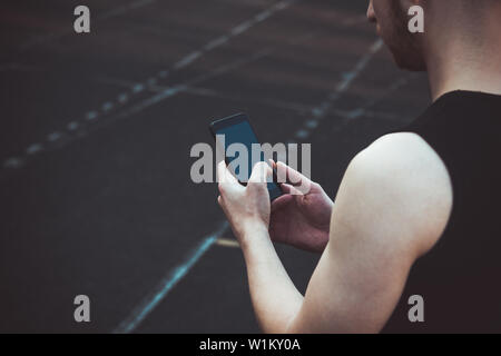 Ein junger Mann in schwarzer Kleidung am Telefon sucht. fitness Athleten auf dem Sportplatz. Training mit Gadgets. Warm up Körper Vorbereitung für die Summe Stockfoto