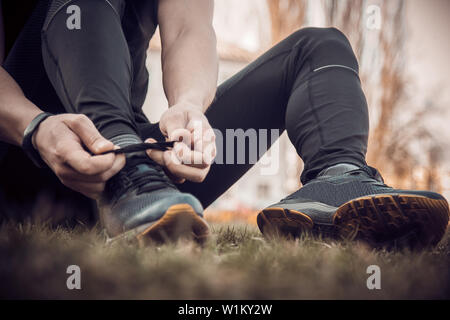 Ein junger Mann in schwarzer Kleidung ist das Binden der Schnürsenkel auf der Sneakers hautnah. fitness Sportler sitzen auf dem Sportplatz auf dem Gras. Warm up Körper Vorbereitung Stockfoto