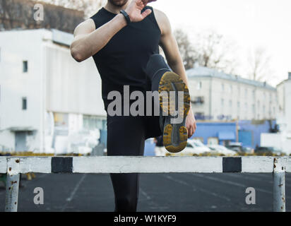 Ein Mann in schwarzer Kleidung ist Training im Freien mit einer Barriere. fitness Athleten auf dem Sportplatz. Training mit Hürde. Warm up stretching Beine. Körper Stockfoto