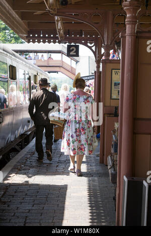 Kidderminster, Großbritannien. Juni 2019. Severn Valley Railways "die Rückkehr in die 40er Jahre" beginnt diesen Sommer mit einem fabelhaften Start in die Zeit mit kostümierten Reenactors, die eine authentische Nachbildung des Zweiten Weltkriegs in Großbritannien bieten. Von hinten sieht man ein Paar aus den 1940er Jahren, das die Bahnsteige eines historischen Bahnhofs entlang an einem alten Wagen der wartenden Dampfeisenbahn vorbeigeht. Bild: Lee Hudson Stockfoto