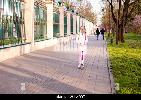 Lustige blonde Mädchen lernen auf Weiß scooter im warmen grünen Park in der Mitte der großen Stadt zu skaten. Sommer aktivitäten Konzept. Stockfoto