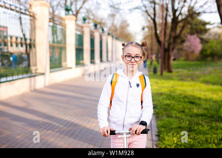 Lustige blonde Mädchen lernen auf Weiß scooter im warmen grünen Park in der Mitte der großen Stadt zu skaten. Sommer aktivitäten Konzept. Stockfoto